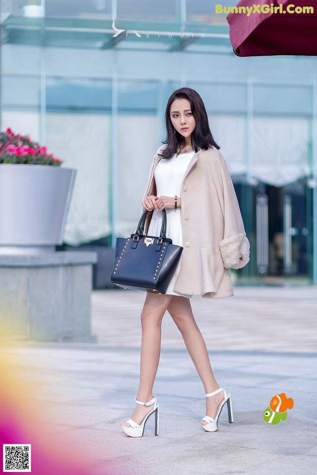 A woman in a white dress and a beige coat is walking down the street.